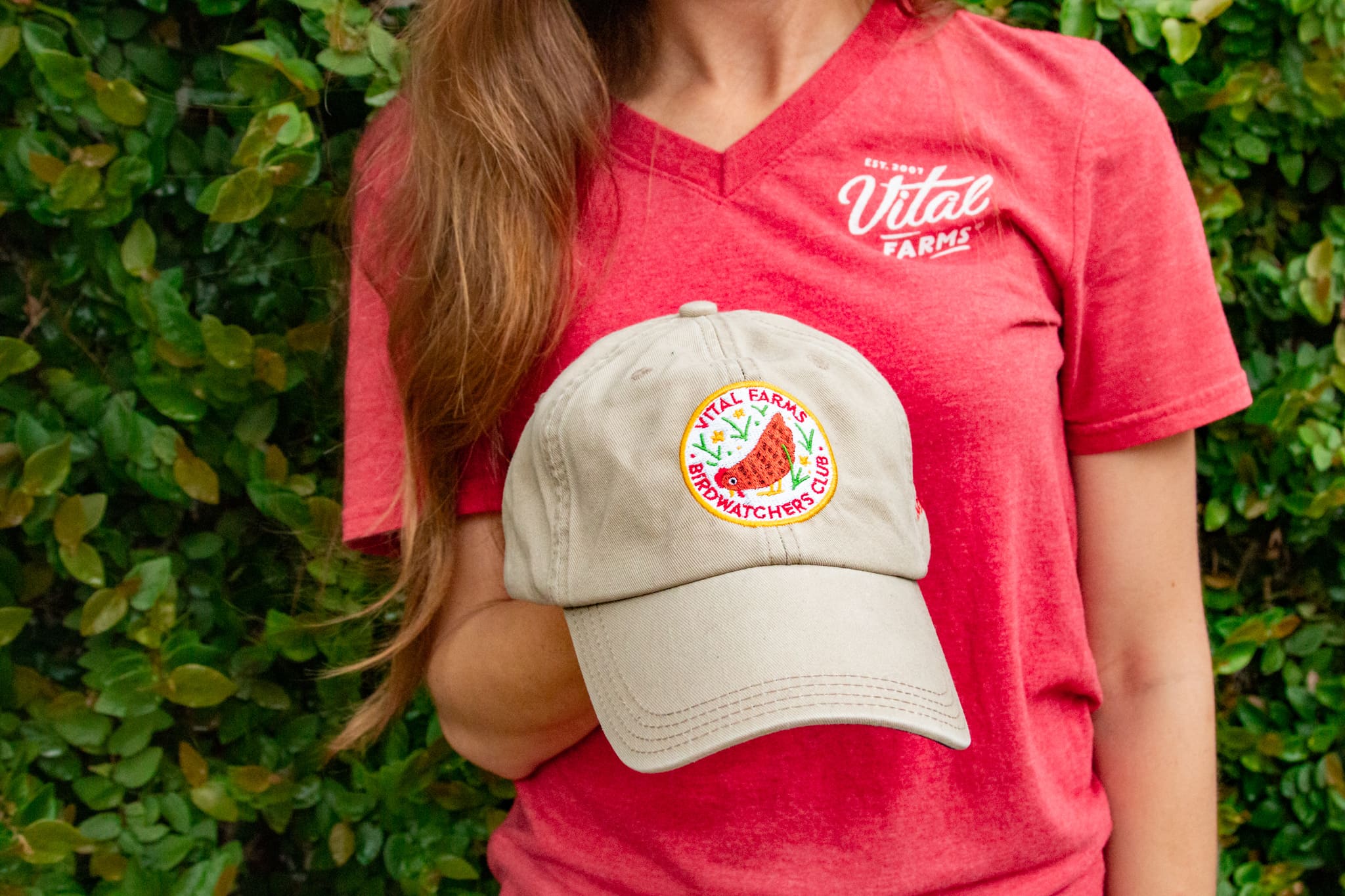 A woman in a red t-shirt with a vital farms logo holding a vital farms birdwatchers baseball cap.